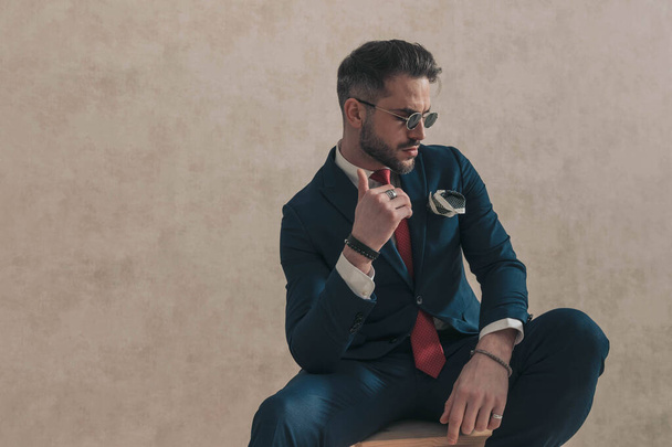 cool bearded businessman in suit holding elbow on knee and looking down while posing in front of beige background - Photo, Image