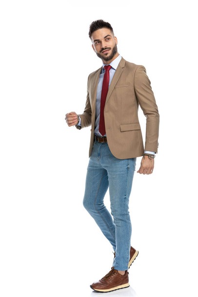 Ganzkörperbild eines lächelnden Mannes in schickem lässigem Outfit, der wegschaut und auf weißem Hintergrund im Studio spaziert - Foto, Bild