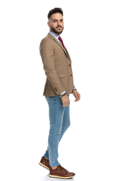 vista lateral de homem morena bonito em roupa casual inteligente andando e posando na frente de fundo branco no estúdio - Foto, Imagem
