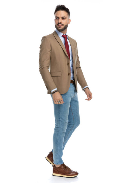 full body picture of sexy unshaved guy with red tie and blue jeans looking away and confidently posing in front of white background in studio - Photo, Image