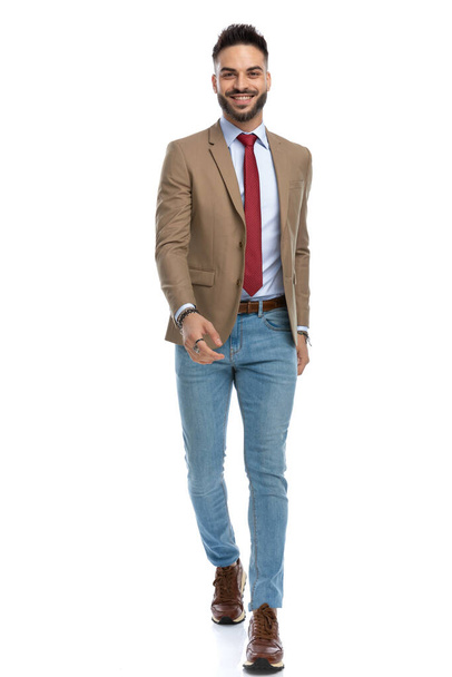 foto del tipo feliz con pantalones vaqueros azules con zapatillas de deporte en un traje casual inteligente caminando en el estudio y sonriendo frente al fondo blanco - Foto, imagen