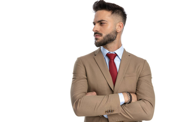 confiant homme d'affaires non rasé pliant les bras et regardant de côté tout en posant devant fond blanc en studio - Photo, image