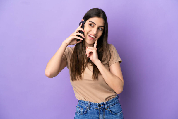 Jeune femme caucasienne utilisant un téléphone mobile isolé sur fond violet penser une idée tout en levant les yeux - Photo, image