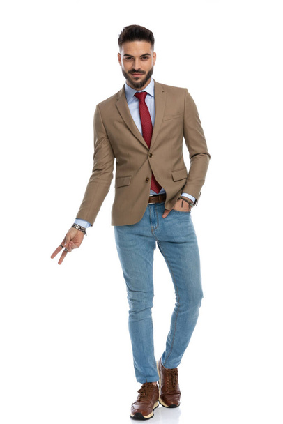 charming young guy with red tie smiling and making peace sign with one hand, holding the other one in pocket and walking on white background - Foto, Imagen