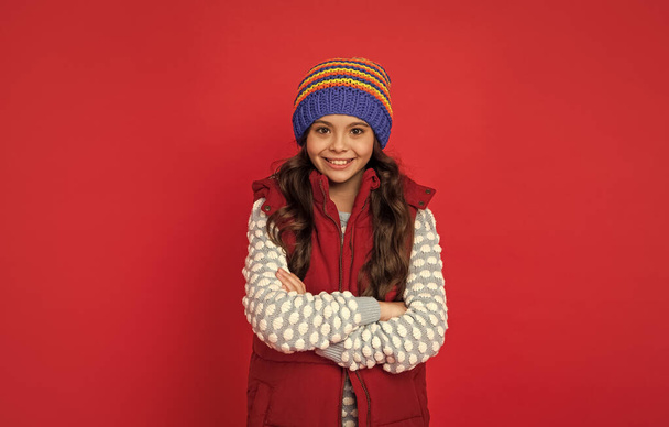 cheerful child in knitted winter hat and down vest crossed hands on red background, cold season. - Foto, immagini