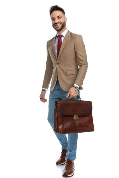 full body picture of happy young man with red tie holding suitcase and smiling while posing in front of white background in studio - Фото, изображение