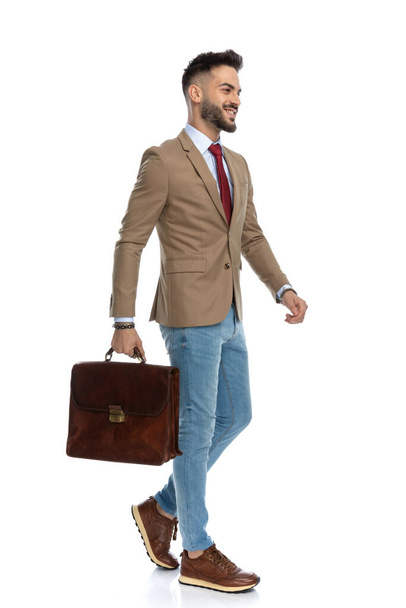 full length picture of proud businessman with suitcase wearing blue jeans and sneakers looking to side and smiling on white background in studio - Foto, imagen
