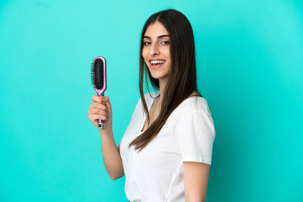 Young caucasian woman with hair comb isolated on blue background smiling a lot - Foto, Imagem