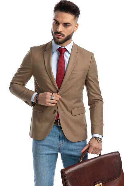 bearded young businessman with suitcase unbuttoning jacket and looking away while posing on white background - Photo, Image
