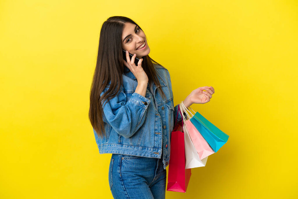 Jonge blanke vrouw geïsoleerd op blauwe achtergrond met winkeltassen en bellen naar een vriend met haar mobiele telefoon - Foto, afbeelding