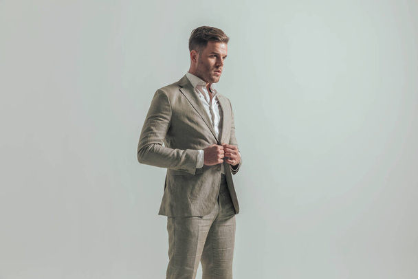 portrét pohledného podnikatele hledajícího na stranu při rozepínání obleku a pózování před šedým pozadím ve studiu - Fotografie, Obrázek