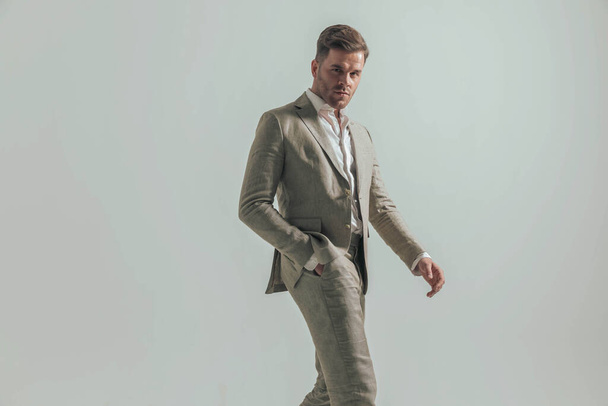 πλαϊνή άποψη του σέξι άνδρα με ανοιχτό γιακά πουκάμισο κρατώντας το χέρι στην τσέπη και περπατώντας μπροστά από το γκρι φόντο στο στούντιο - Φωτογραφία, εικόνα