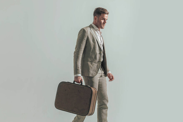 side view of handsome stylish man with baggage looking to side and walking in front of grey background in studio - Photo, Image