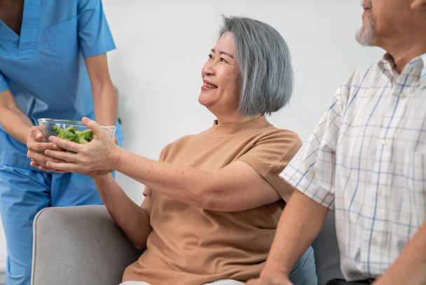 A female nurse serves a bowl of salad to a contented senior couple. Health care and medical assistance for the elderly, nursing home for pensioners. - Photo, Image