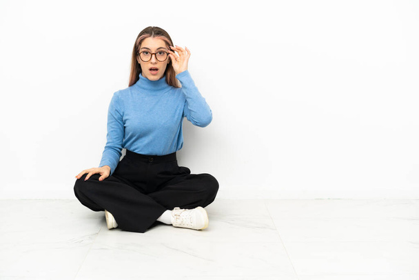 Молода кавказька жінка сидить на підлозі з окулярами і здивована. - Фото, зображення