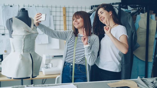 Two cheerful female clothing designers are making funny selfie with smart phone while standing beside clothed mannequin in studio. Attractive women are smiling and posing. - Photo, Image