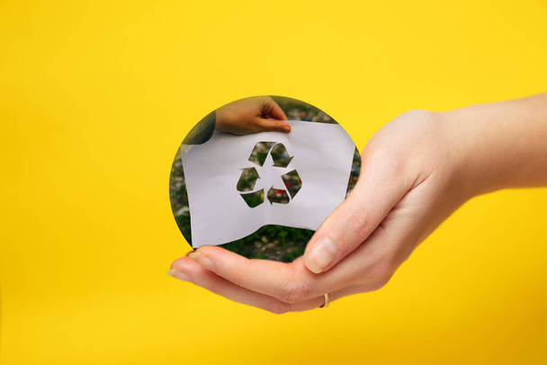Defocus mão segurando papel cortado com o logotipo de reciclagem sobre lixo lixo lixo fundo. Fundo amarelo. Conceito de reciclagem. Poluição do planeta. Fora de foco. - Foto, Imagem