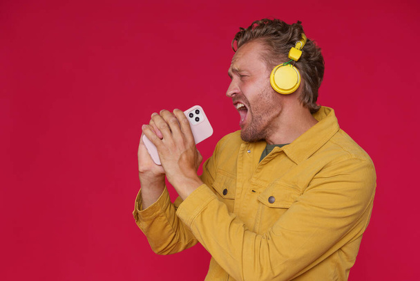 Handsome man 30s singing enjoying his favorite song or track using phone and wireless headphones wearing denim yellow jacket isolated on red background. Joyful man sing while listen music.  - Foto, immagini