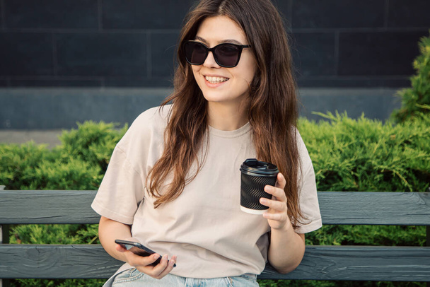 A business young woman in sunglasses sits on a bench with a cup of coffee and a smartphone in her hands. - Fotografie, Obrázek