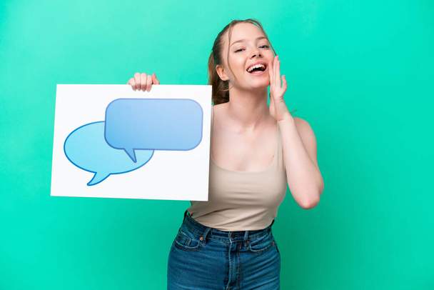Young caucasian woman isolated on green background holding a placard with speech bubble icon and shouting - Photo, Image