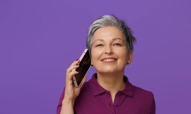 Happy smiling grey haired mature woman talking on the phone. Charming senior woman in purple polo shirt use modern smartphone to communicate happy looking in camera isolated on purple background.  - Φωτογραφία, εικόνα