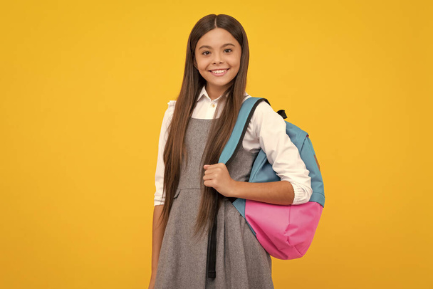 Iskolás lány egyenruhában, iskolatáskával. Iskoláskorú tinédzser tartsa hátizsák sárga elszigetelt háttér. Boldog lány arc, pozitív és mosolygós érzelmek - Fotó, kép