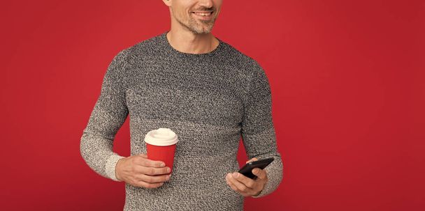 gelukkig knappe man dragen trui hold telefoon en koffiekop, communicatie. - Foto, afbeelding