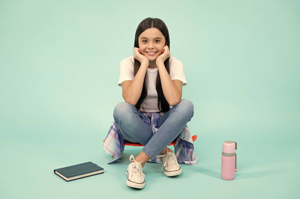 Fashion teen girl wearing trendy t-shirt and jeans sit on skateboard, posing on blue background. Fashion teenager, casual look. Happy teenager, positive and smiling emotions of teen girl - Foto, Imagem