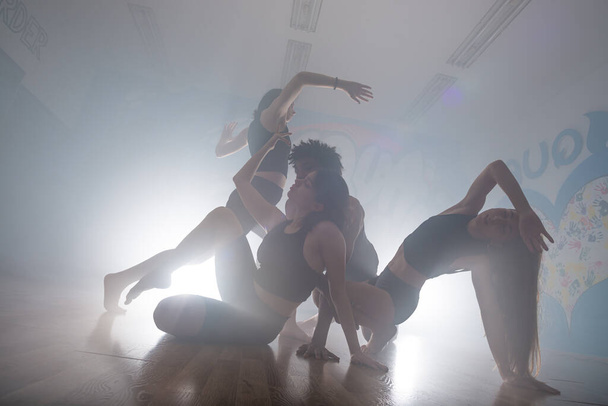 Ομάδα από πολυφυλετικές χορεύτριες που απολαμβάνουν funky hip hop κινήσεις σε σκοτεινό στούντιο με καπνό και φωτισμό. Ομάδα νεαρών χορευτών χιπ-χοπ που παίζουν στη σκηνή. Ευτυχισμένες χορεύτριες. - Φωτογραφία, εικόνα