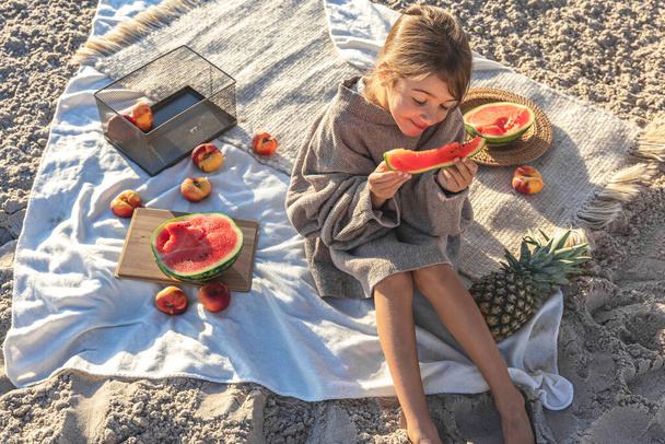 A little girl on a sandy seashore on a blanket at a picnic with fruits eats watermelon. - Foto, afbeelding