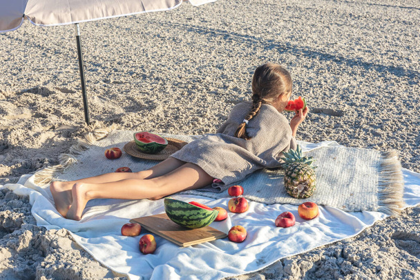 Little funny girl lies on a blanket on the sea beach, rest by the sea with fruits. - Foto, Imagen