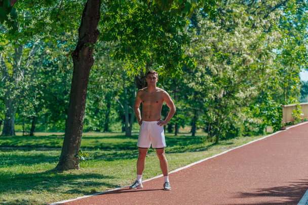 Mladý sportovec bez trička stojí na běžecké dráze v parku - Fotografie, Obrázek
