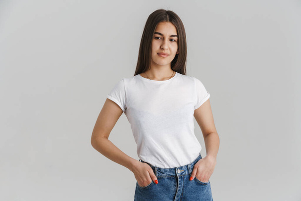 Portrait of a young casual white woman in t-shirt with long brunette hair standing over gray wall background hands in pockets - Foto, Imagem