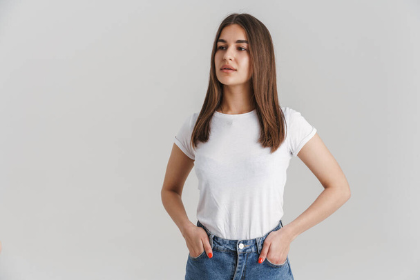 Portrait of a young casual white woman in t-shirt with long brunette hair standing over gray wall background hands in pockets looking aside - Фото, изображение