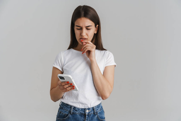 Portrait of a frowning young casual white woman in t-shirt with long brunette hair standing over gray wall background holding mobile phone - 写真・画像
