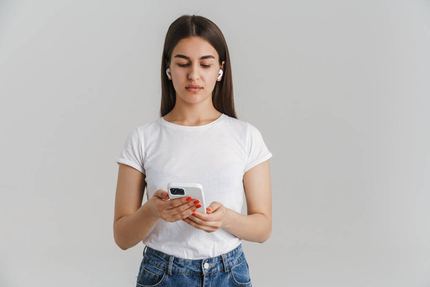 Retrato de una joven mujer blanca casual en camiseta con el pelo largo morena de pie sobre fondo gris de la pared sosteniendo el teléfono móvil escuchando música con auriculares inalámbricos - Foto, imagen