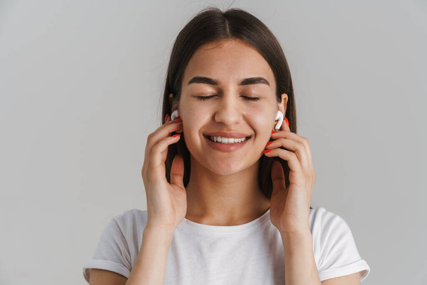 Portrait of a young happy casual white woman in t-shirt with long brunette hair standing over gray wall background listening to music with wireless earphones - Foto, afbeelding