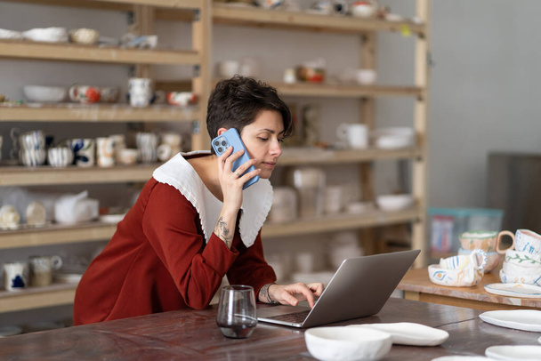 Young creative woman artisan artist ordering materials for ceramic work by phone while working in studio or workshop, female pottery shop owner talking with client via cellphone and typing on laptop - 写真・画像