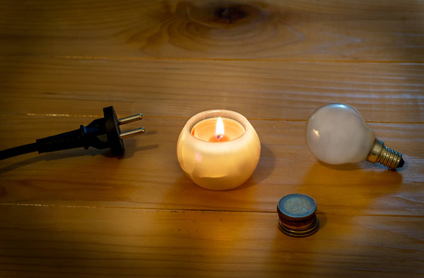 Sähkön säästäminen. Sähkölamppu ja kaapeli pistoke kynttilä puinen pöytä. Eugo-kolikoita. Menneisyyteen paluun symboli. Hehkulamppu ja kynttilä. Sähkökatko tai sähkökatkos. Korkeat sähkön hinnat. - Valokuva, kuva