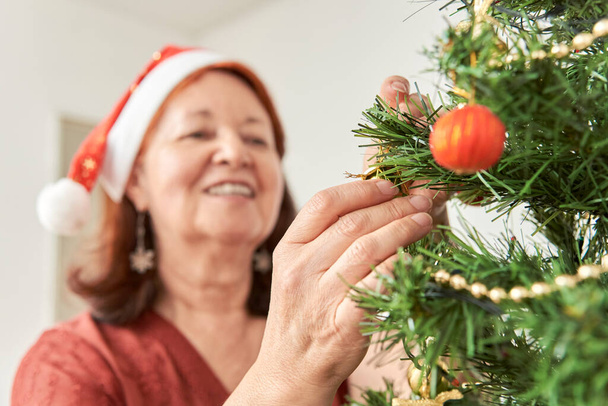 Gelukkige oudere Spaanse vrouw glimlachend terwijl ze een kerstboom versiert met een rode kerstmuts. Selectieve focus op haar handen. De vreugde van de vakantie. - Foto, afbeelding