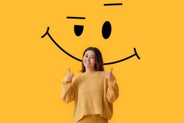 Hermosa joven asiática mostrando el pulgar hacia arriba y dibujado sonrisa sobre fondo amarillo - Foto, Imagen