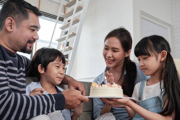 Happy Asian Thai family, young son is surprised with birthday cake, blows out candle, and celebrates joy party with parents and sister in living room together, wellbeing domestic home special event. - Foto, Bild