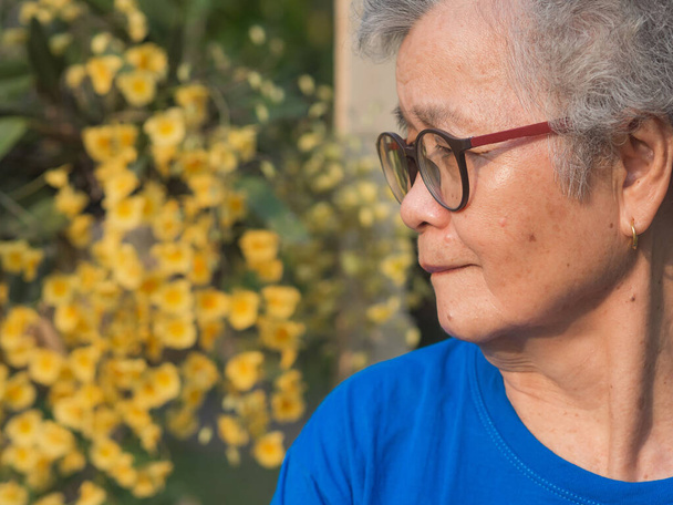 Старшая женщина с короткими седыми волосами в очках, улыбается и смотрит на желтые орхидеи, стоя в саду. Вид сбоку. Пространство для текста. Концепция пожилых людей и здравоохранения. - Фото, изображение