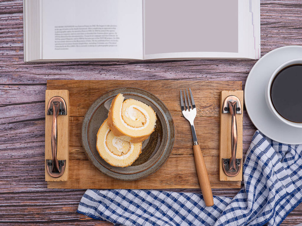 Κάτοψη βιβλίου με roll cake σε πιάτο σε ξύλινο δίσκο και λευκό φλιτζάνι καφέ πάνω σε ξύλινο τραπέζι. Χώρος για κείμενο. Έννοια της χαλάρωσης. - Φωτογραφία, εικόνα