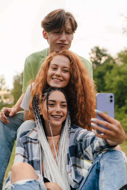 Τρεις χαρούμενοι χίπστερ φίλοι διασκεδάζουν στο πάρκο, βγάζουν μια σέλφι, απολαμβάνουν τη στιγμή. Άτομα που χρησιμοποιούν smartphone για τη χρήση της ιστοσελίδας κοινωνικής δικτύωσης. Πόλη - Φωτογραφία, εικόνα