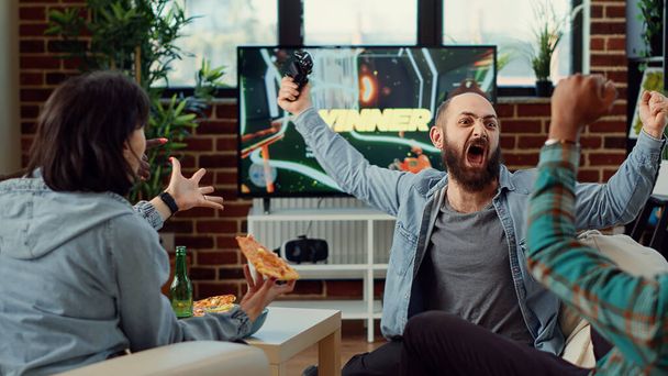 Group of friends feeling happy about winning gaming competition, playing video games online on tv console. People enjoying shooting challenge on television, having fun at home. - Photo, Image