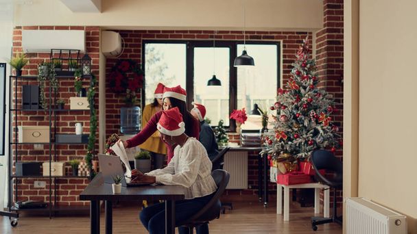 Diverse Leute arbeiten in festlichen Büros, planen Berichte und tragen Weihnachtsmänner. In der Winterferienzeit mit Weihnachtsbaum auf Hochtouren arbeiten. Zeitraffer. - Foto, Bild