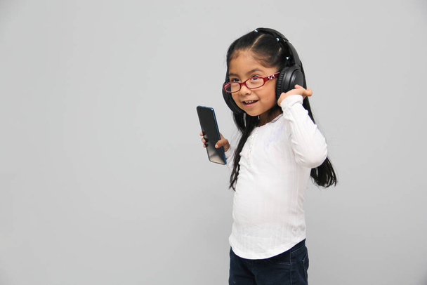 4-year-old brunette Latin girl with glasses listens to music in her headphones connected to her cell phone dances, sings and enjoys the rhythm happy and excited - Fotografie, Obrázek