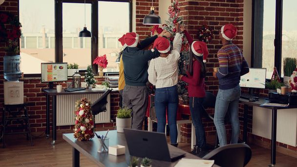 スタートアップオフィスでクリスマスツリーを飾る同僚の多様なグループは、冬の季節の装飾品で幸せとお祝いを感じています。光と装飾を使用してクリスマスを祝う｜eve holiday. - 写真・画像
