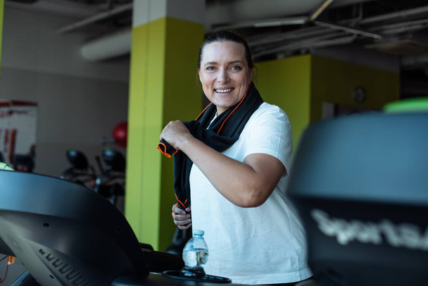 Happy overweight woman vigorously run on treadmill at fitness gym with towel hanging around neck, side view. Adult plus size person do cardio workout on exercise machine. Training, sport lifestyle. - Photo, Image
